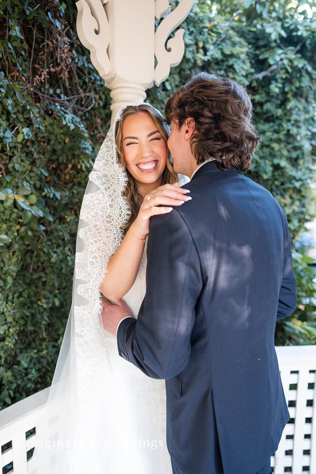 Maya + Max A groom hugging his smiling bride from behind while surrounded by green foliage.
