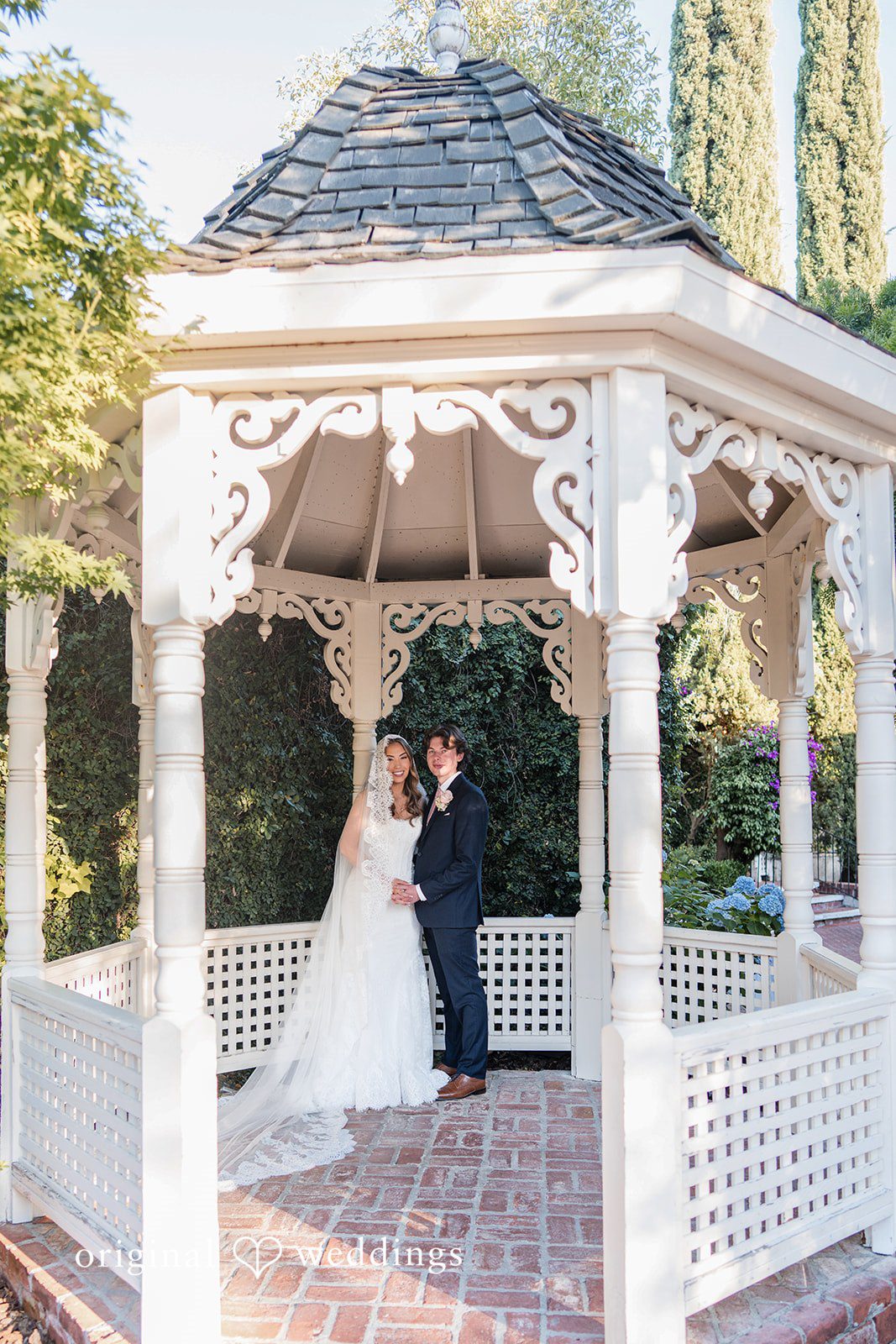 Maya + Max A bride and groom standing together inside a white wooden garden gazebo.