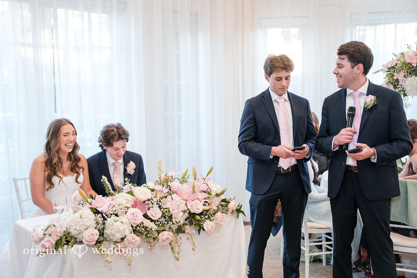 Maya + Max Wedding reception speech with a man speaking into a microphone while the bride and groom laugh at their head table.