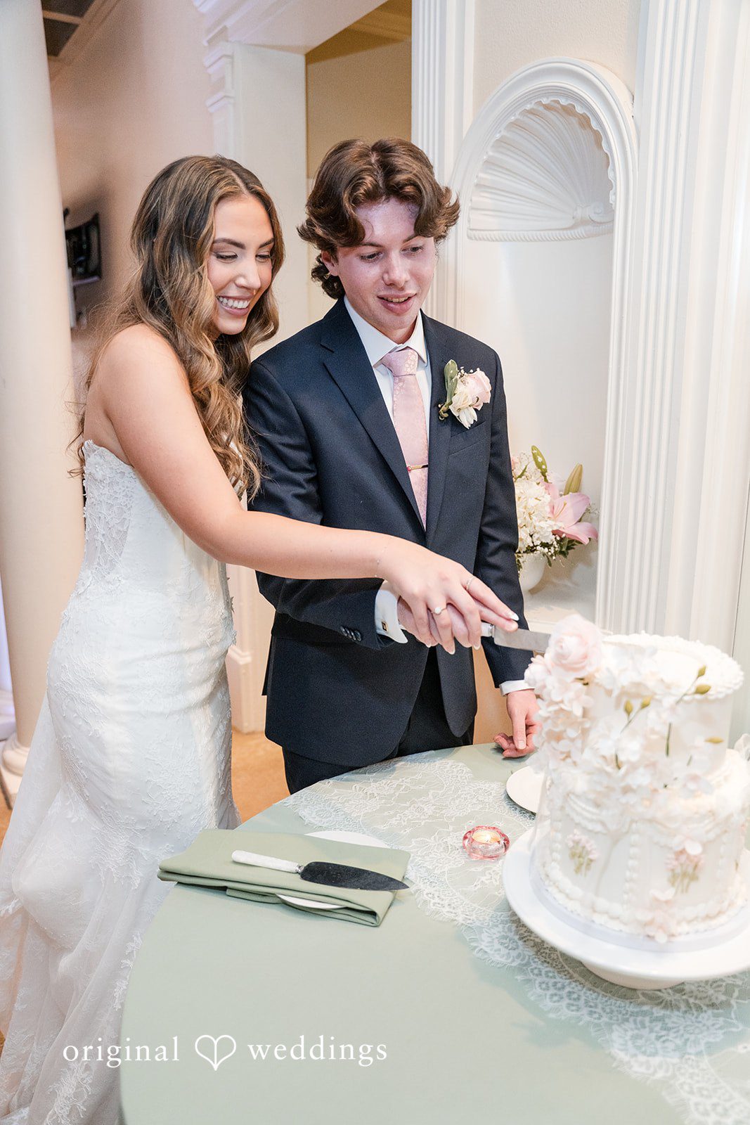 Maya + Max A bride and groom using a silver knife to cut their white wedding cake together.