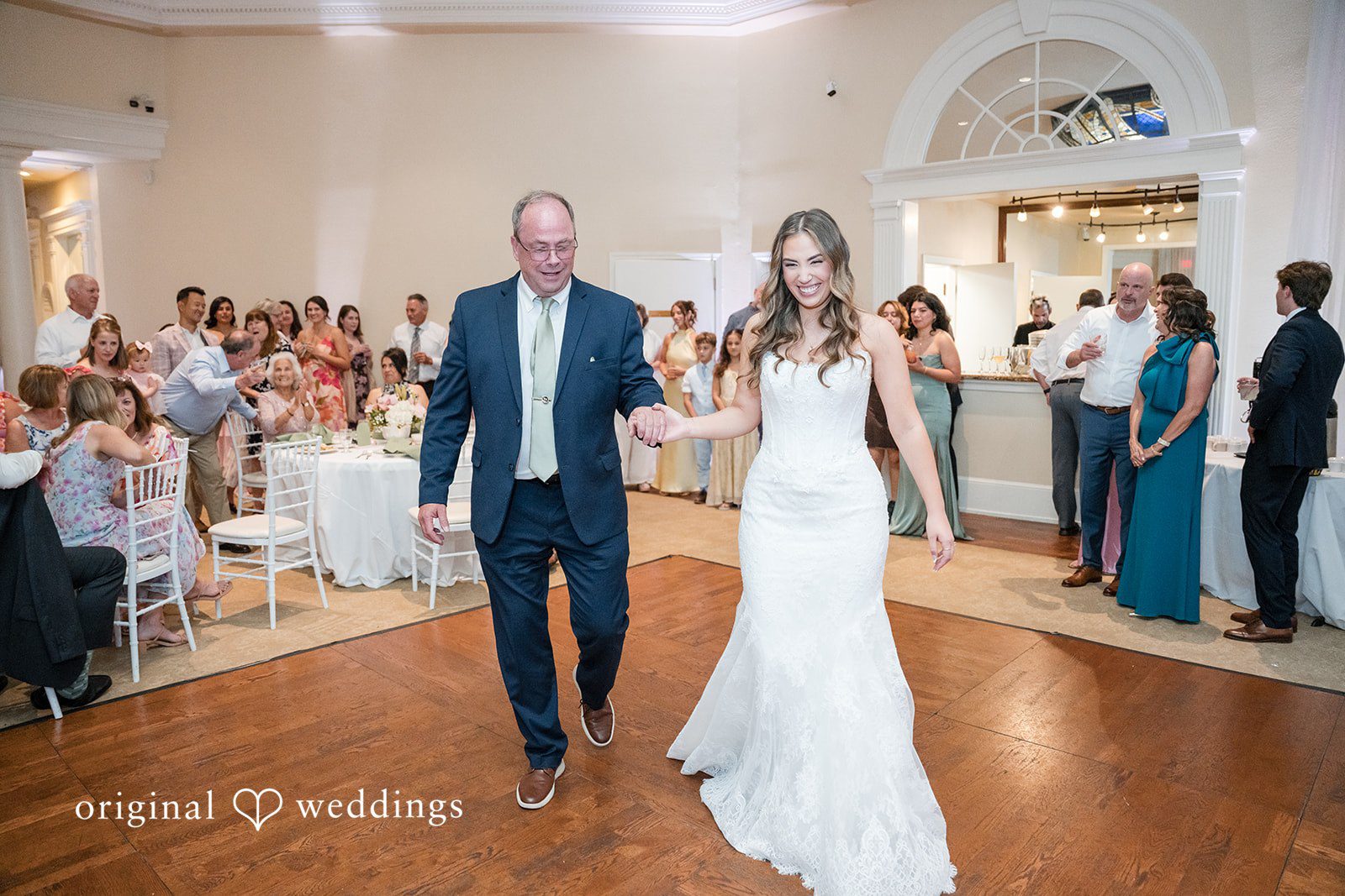 Maya + Max Bride walking onto a wooden dance floor hand-in-hand with a man in a blue suit.