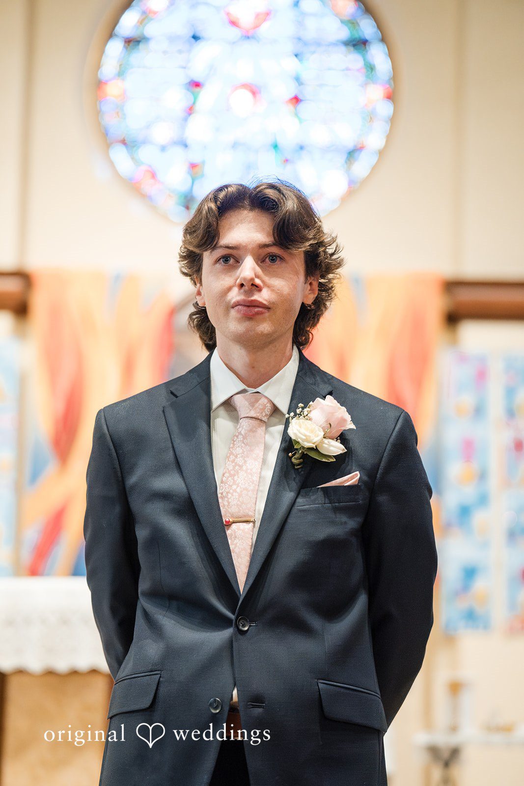 Maya + Max A groom wearing a dark grey suit jacket, a white shirt, and a light pink tie, standing inside a church.