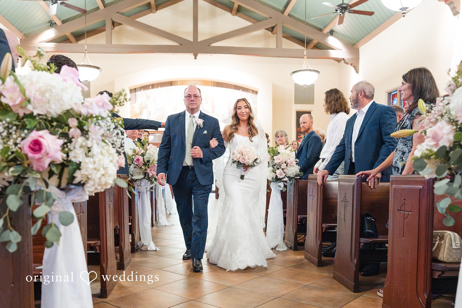 Maya + Max A bride walking down the church aisle on the arm of a man, passed guests sitting in pews during the wedding processional.