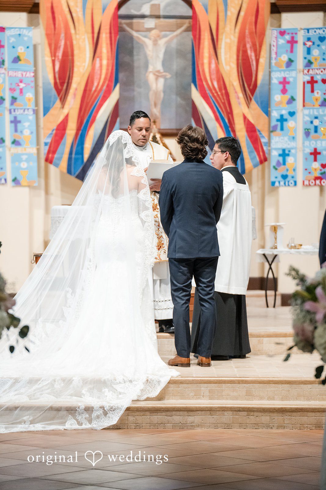 Maya + Max A bride in a long veil and a groom in a dark suit standing before an officiant at a church altar during their ceremony.