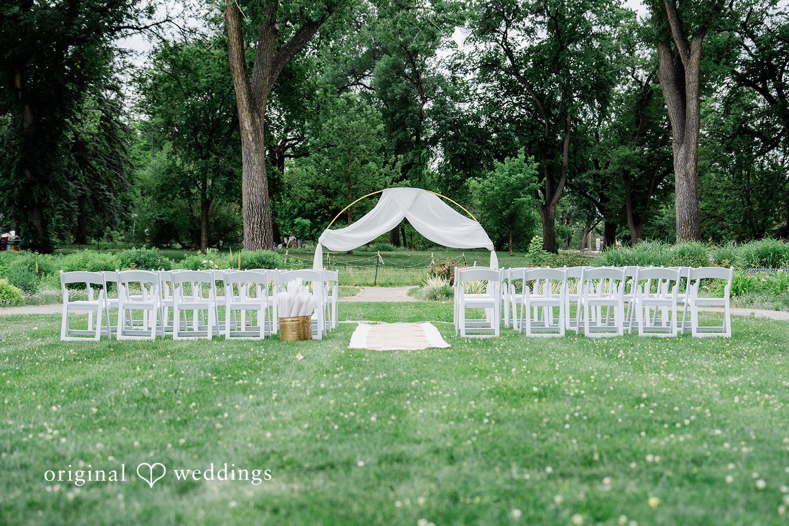 Sophia + Miles White chairs arranged in rows facing a wedding arch draped with white fabric on a lawn.