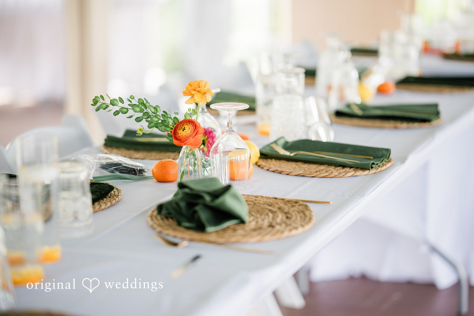 Sophia + Miles Long wedding reception table set with green napkins, white plates, and small vases of orange flowers.