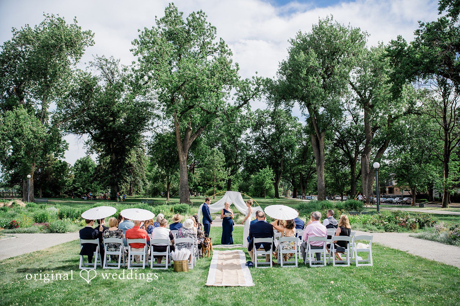Sophia + Miles Distant wide shot of a wedding ceremony in progress with guests seated on a lawn surrounded by trees.