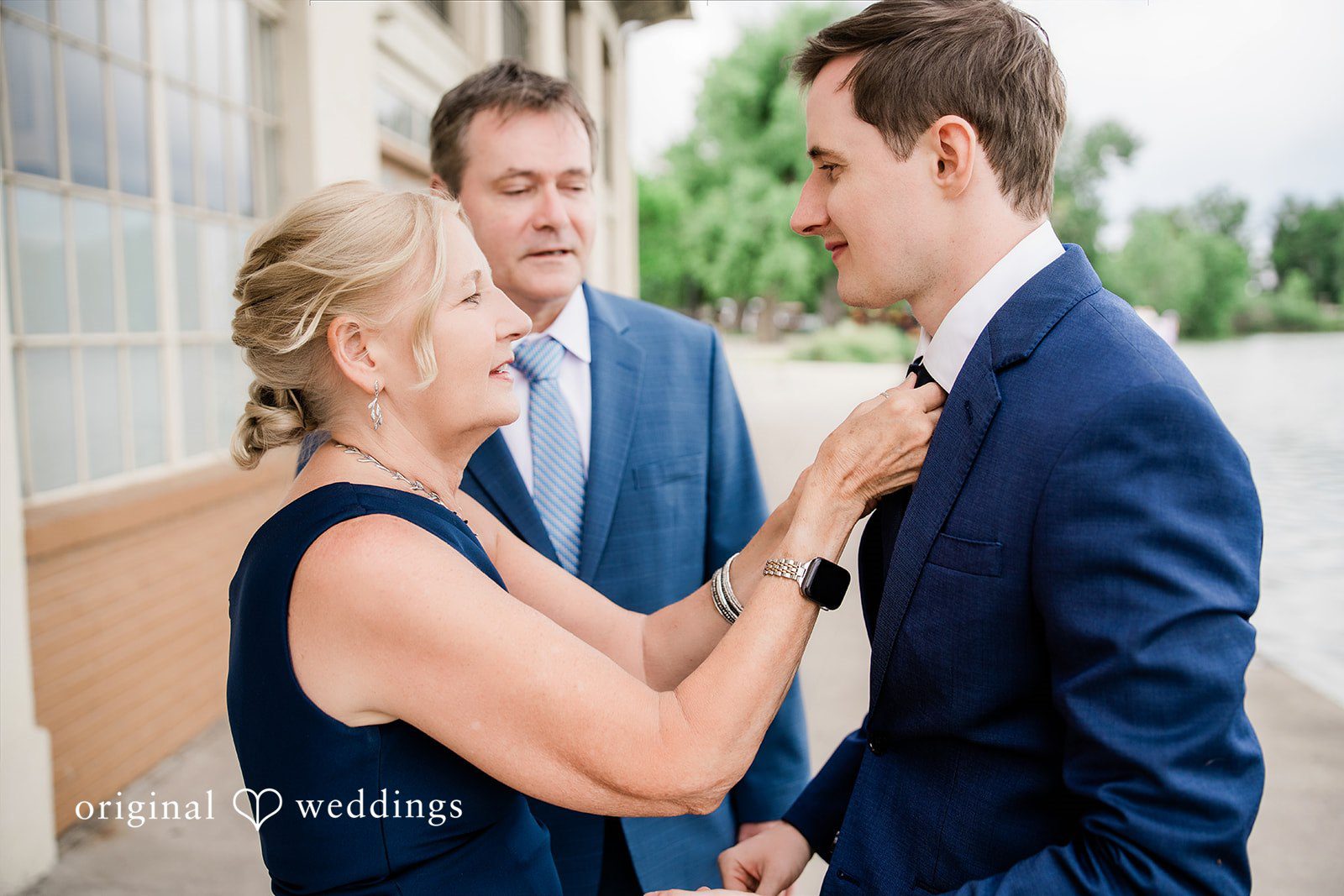 Sophia + Miles A bride smiling while adjusting the boutonniere on the groom's navy blue suit jacket.