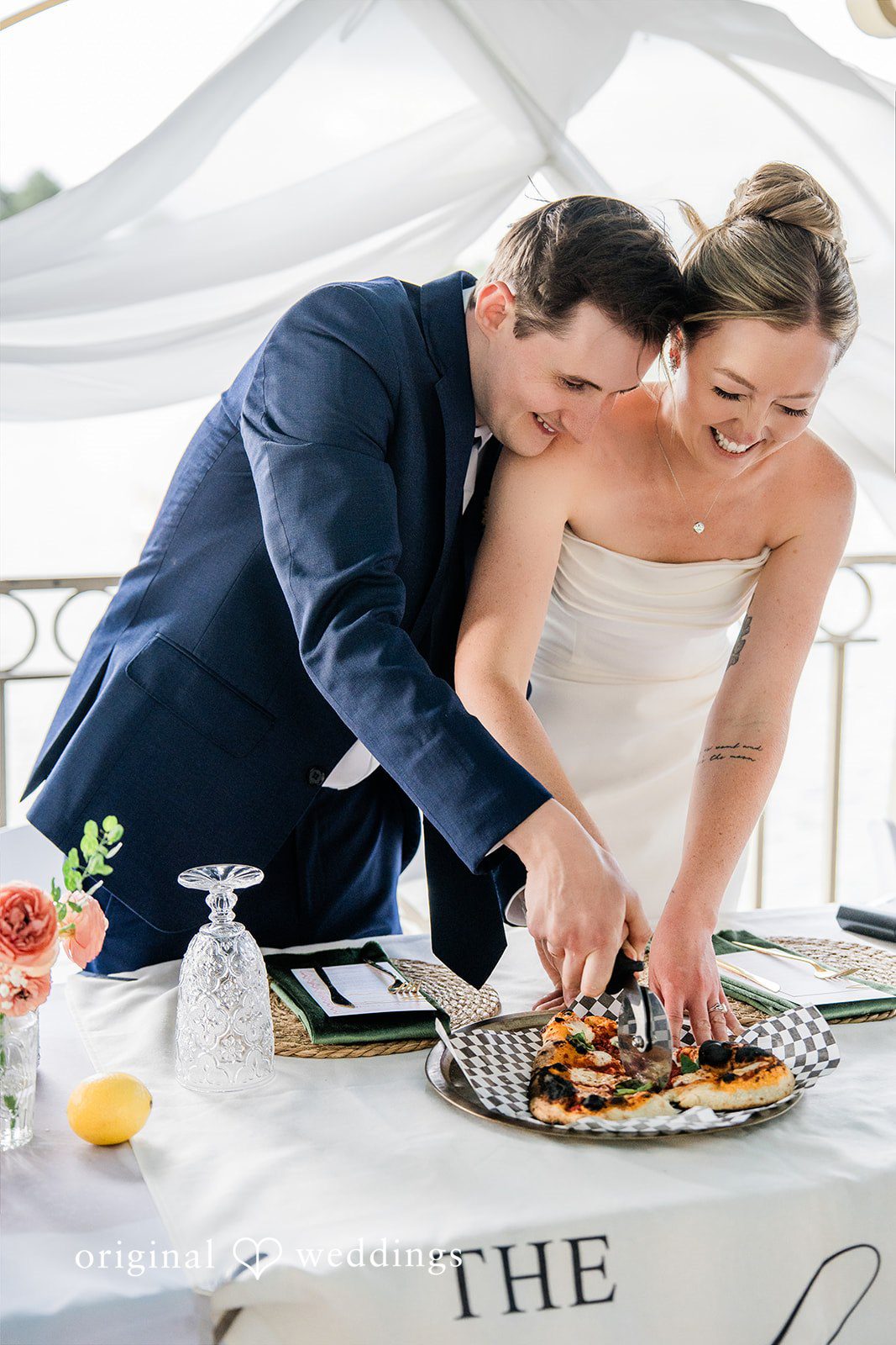 Sophia + Miles A bride and groom smiling while cutting their wedding pizza together at a reception table.