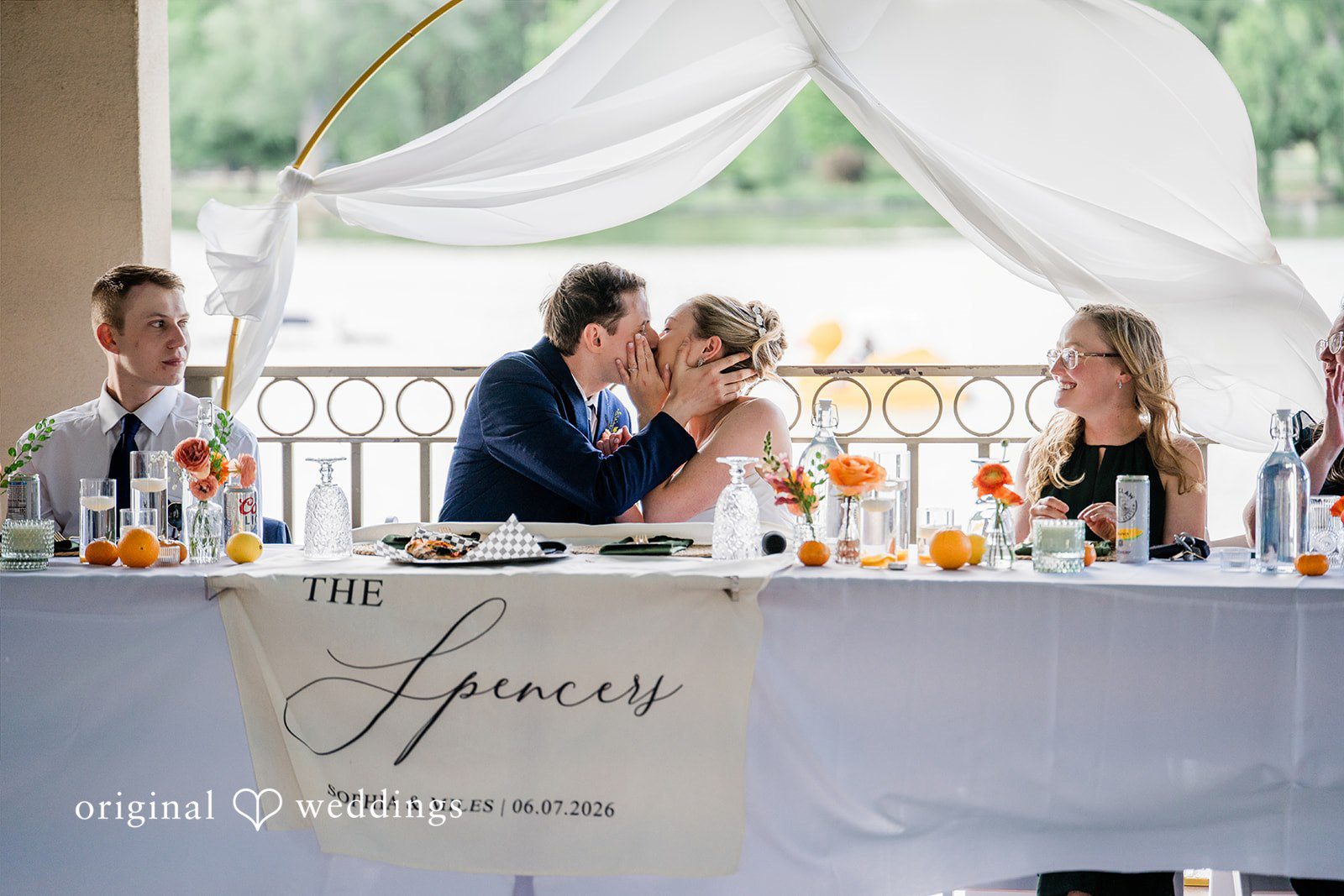 Sophia + Miles A bride and groom kissing at the center of a long head table draped in white fabric.