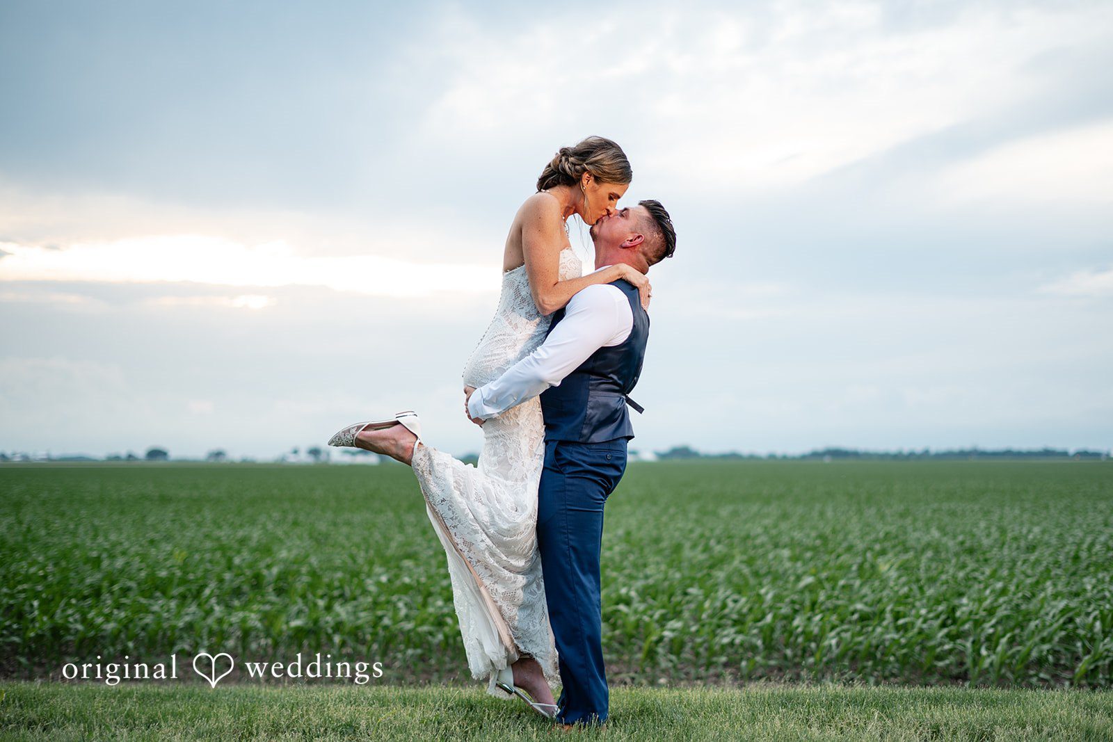 Groom lifting his bride during a romantic kiss beside open countryside fields