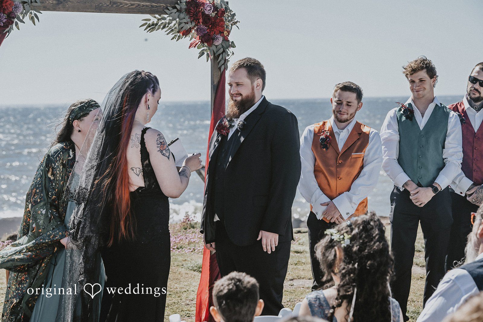 Wedding party in formal dark attire standing together by the coast.