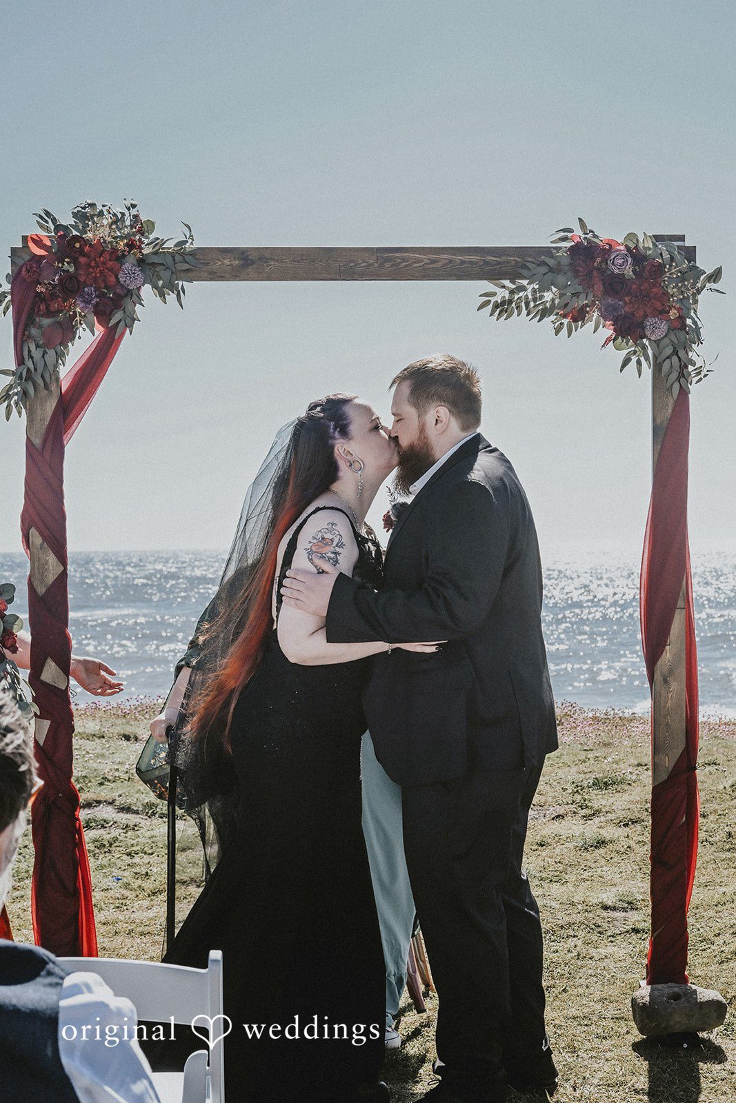 Newlyweds sharing a kiss under a wooden arch next to the ocean.