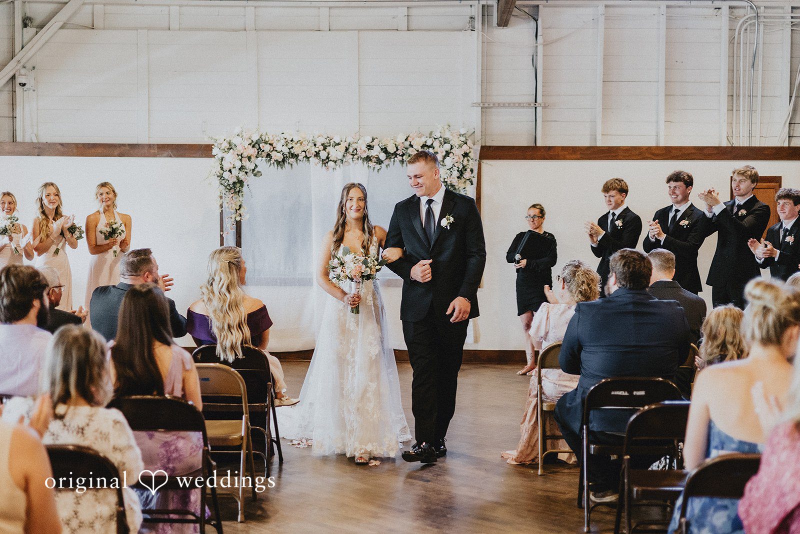 Emily + Ethan A bride and groom walking down the aisle together after their ceremony, smiling as guests applaud.