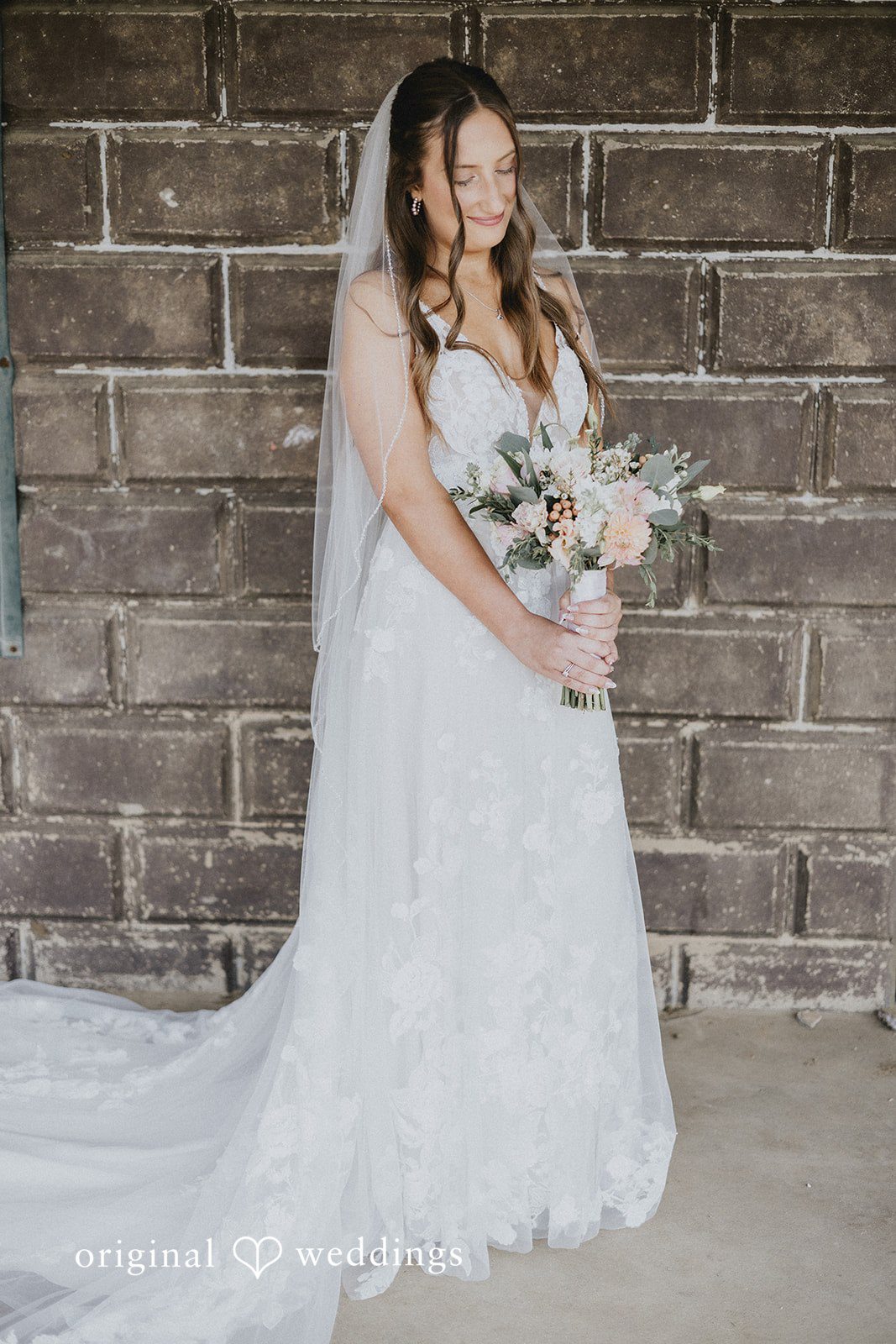 Emily + Ethan Bride in a lace wedding gown standing by a rustic wall.