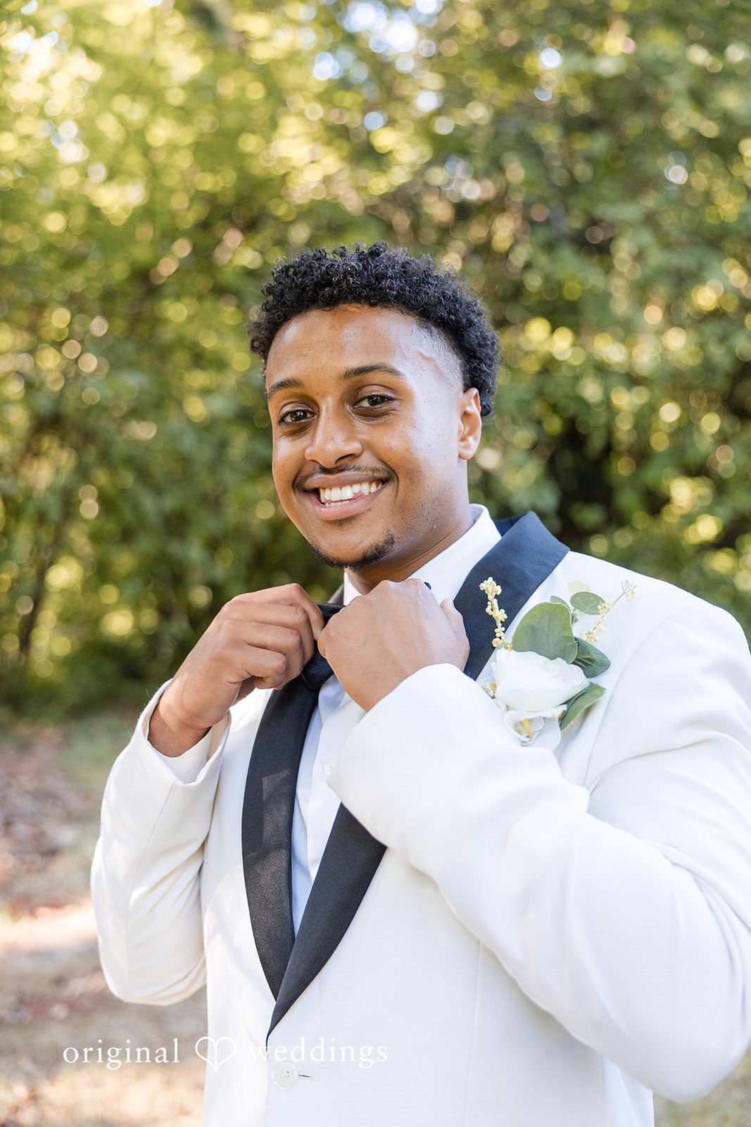 Close-up of a groom adjusting his bowtie in a white tuxedo jacket.