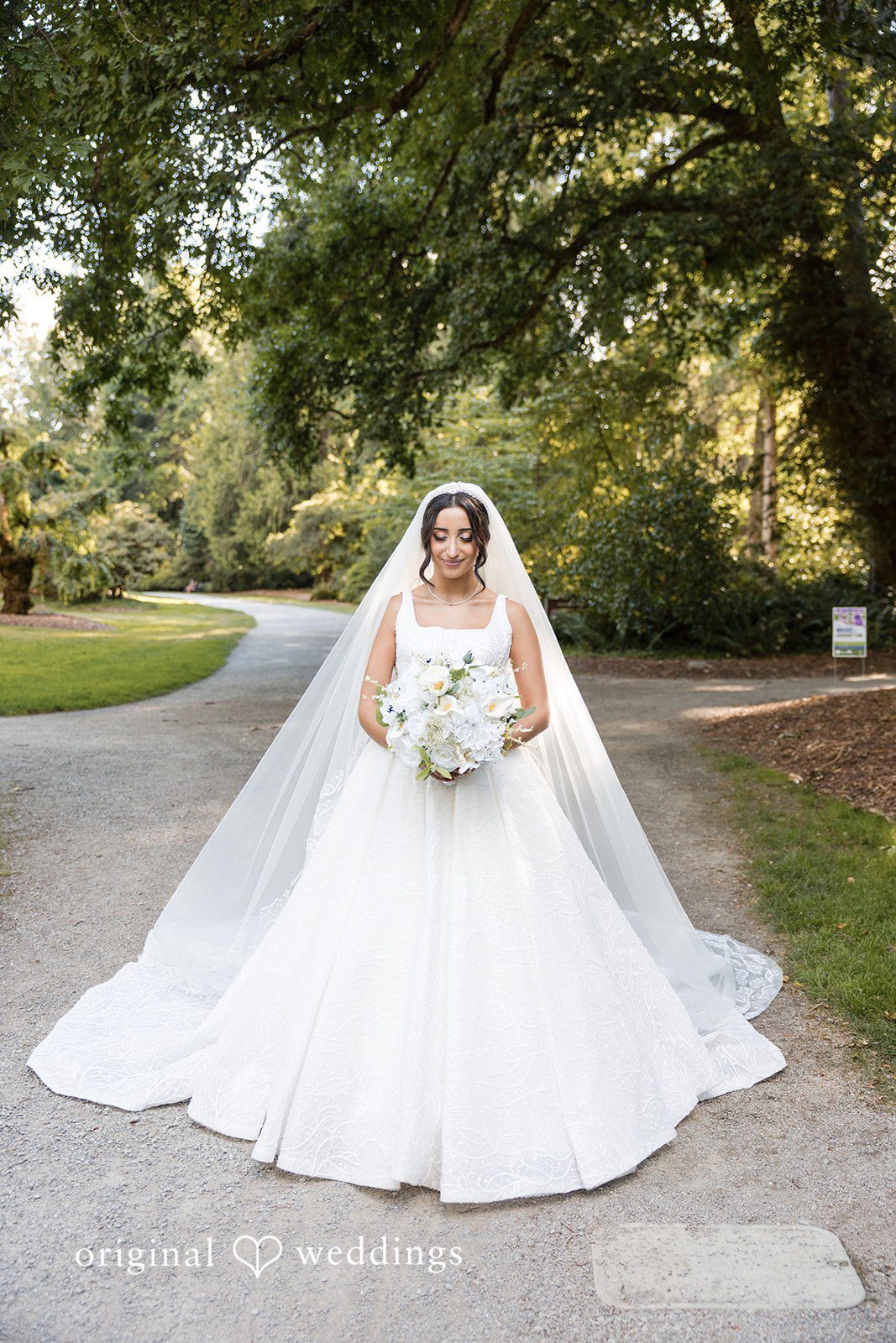 A bride in a full white wedding dress sitting on a pathway.