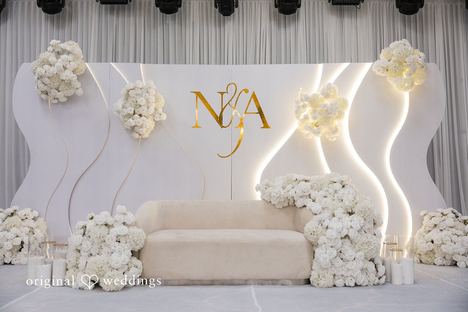 A white wedding reception stage decorated with lights and floral arrangements.