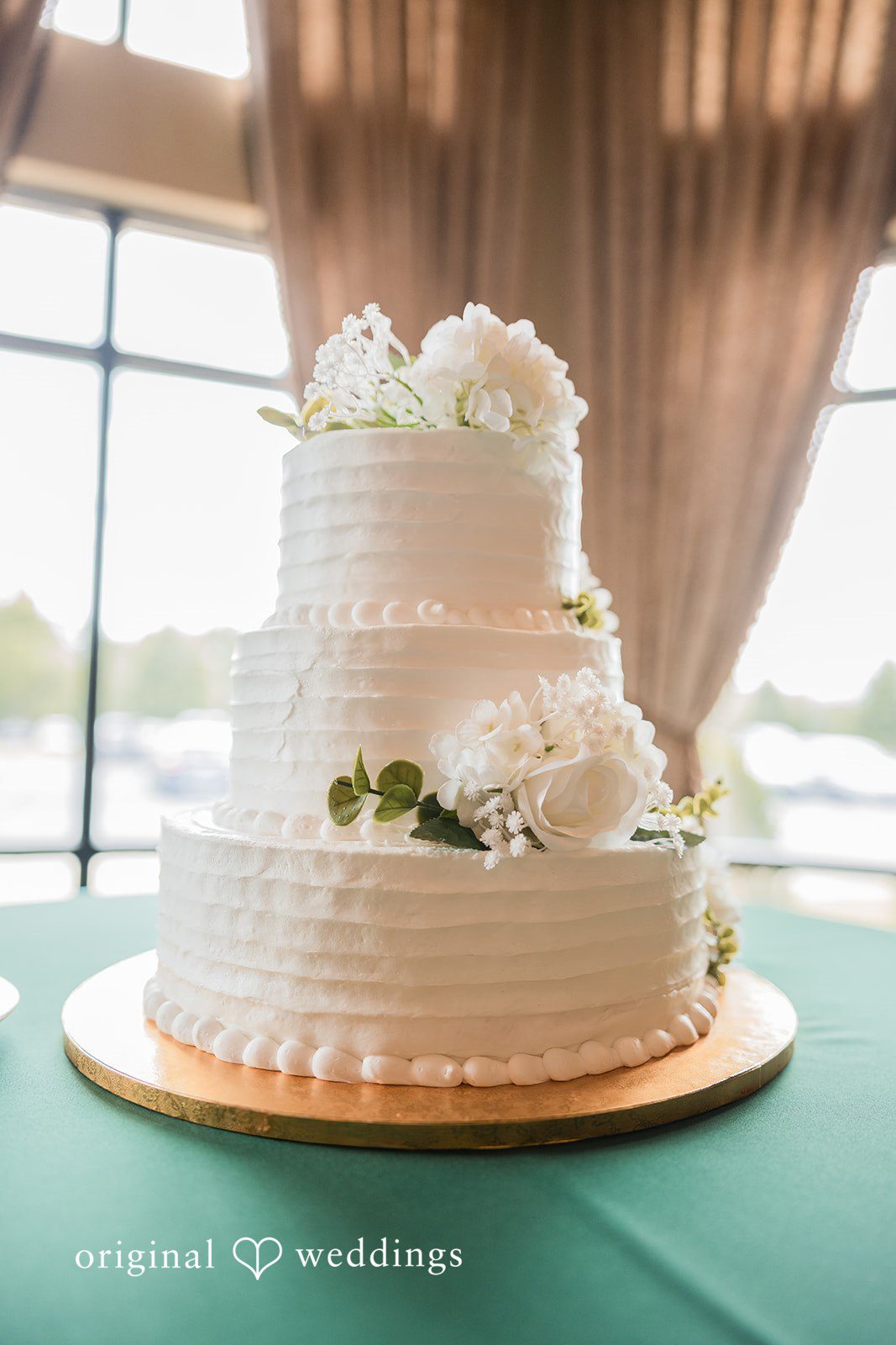 Christopher + Maxine Three-tiered white wedding cake decorated with elegant textures sitting on a green covered table.
