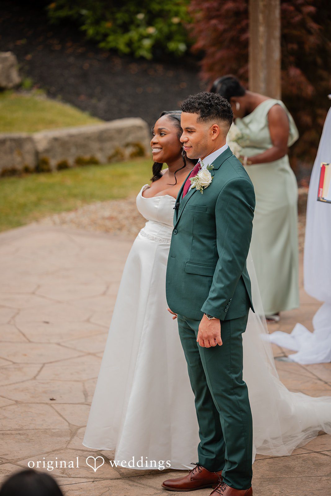 Christopher + Maxine Bride in a white gown and groom in a green suit standing close together on a brick pathway.