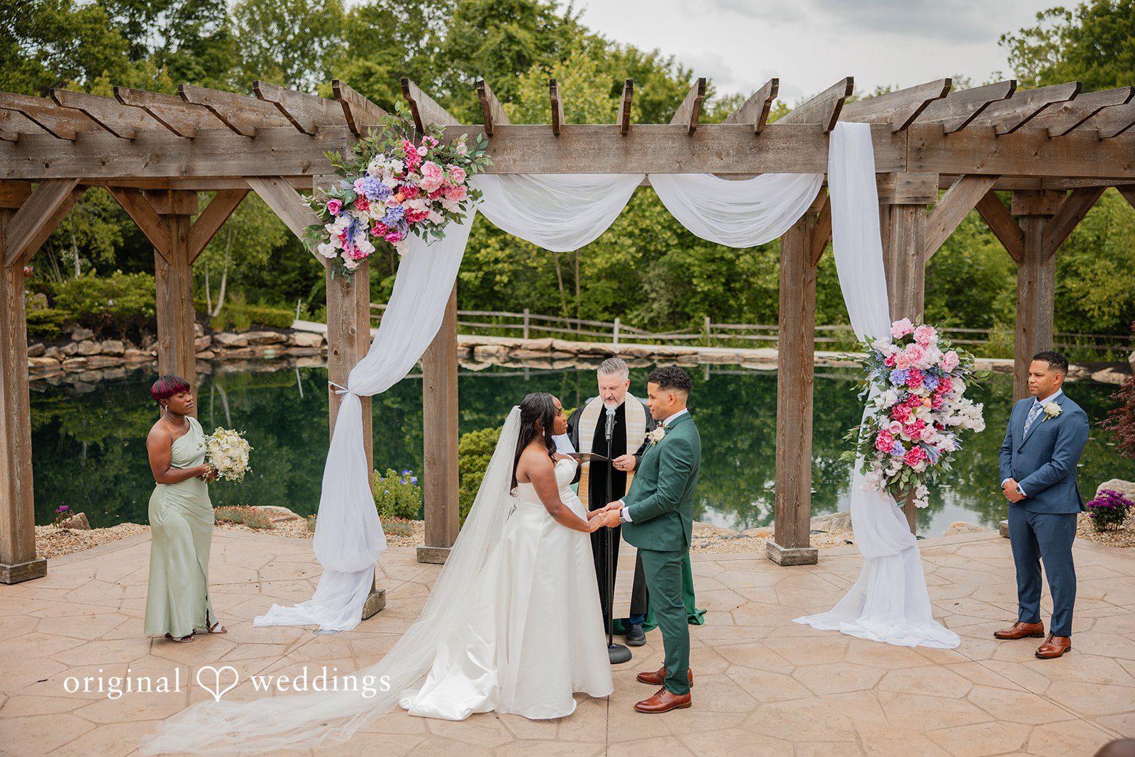 Christopher + Maxine Bride and groom facing each other on a paved path beneath a wooden pergola draped in white fabric.
