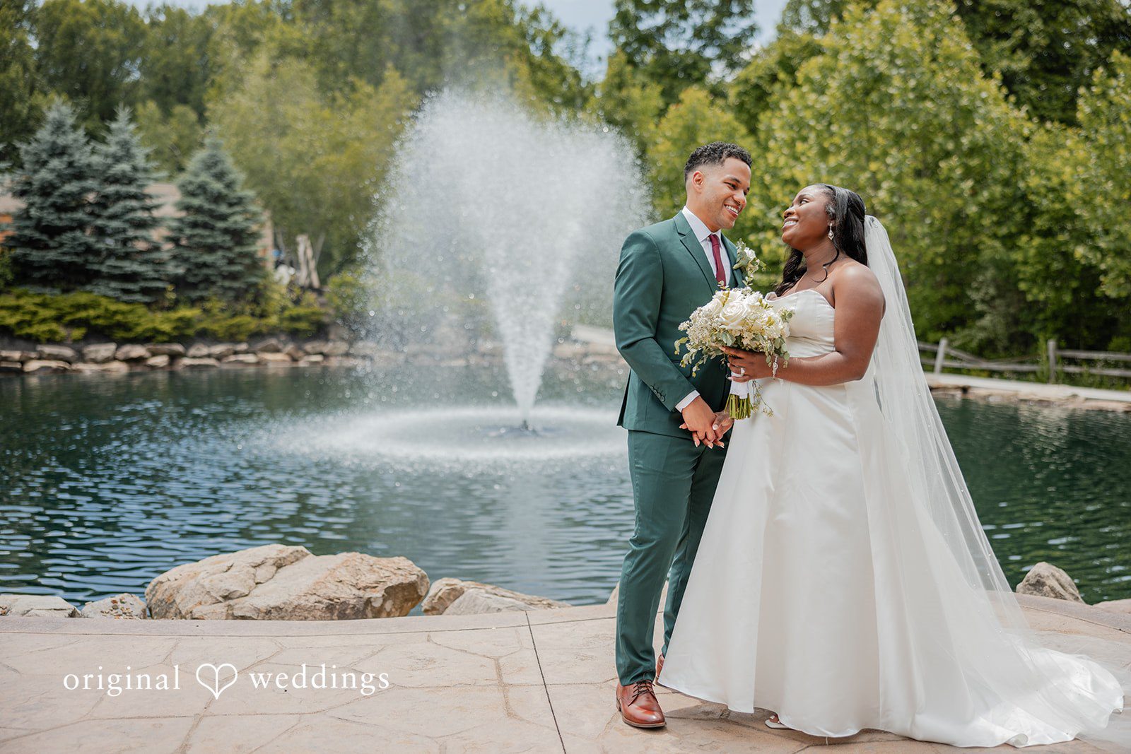 Christopher + Maxine Bride and groom standing near a large pond fountain with a stone wall backdrop.