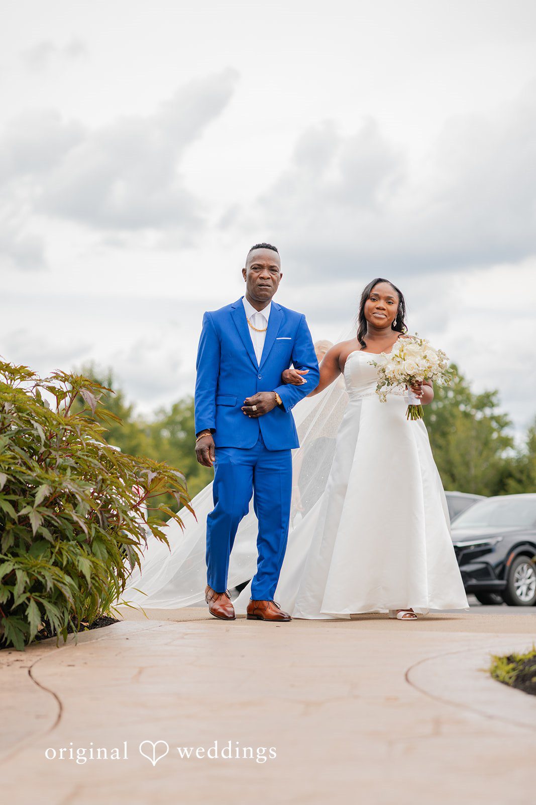 Christopher + Maxine Bride wearing a white gown walking down a pathway alongside a man in a vibrant blue suit.