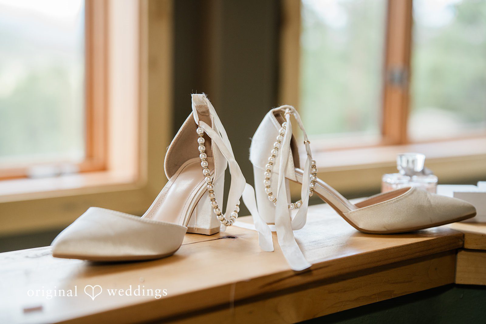 Madelyn + Erin A pair of white high-heeled bridal shoes resting on a wooden window sill.