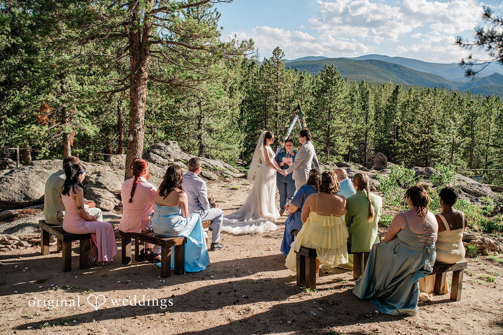Madelyn + Erin Guests seated on benches watching a wedding ceremony take place in the woods.