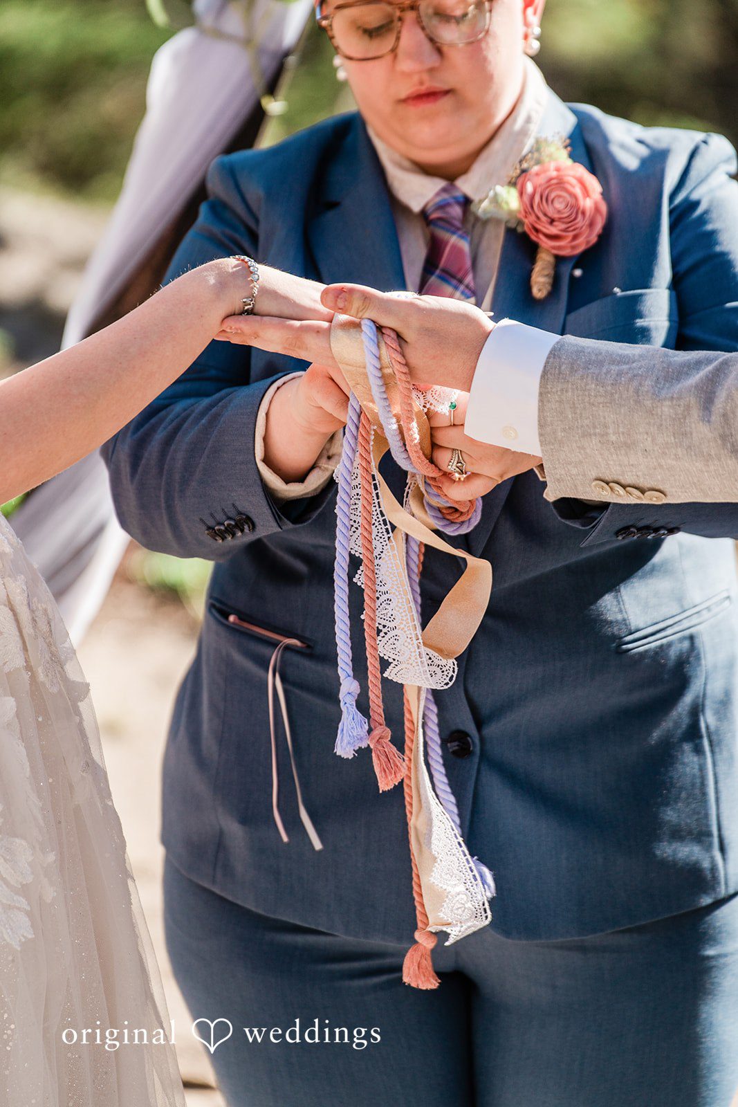 Madelyn + Erin A close-up of a couple's hands during a handfasting ritual with colorful ribbons.
