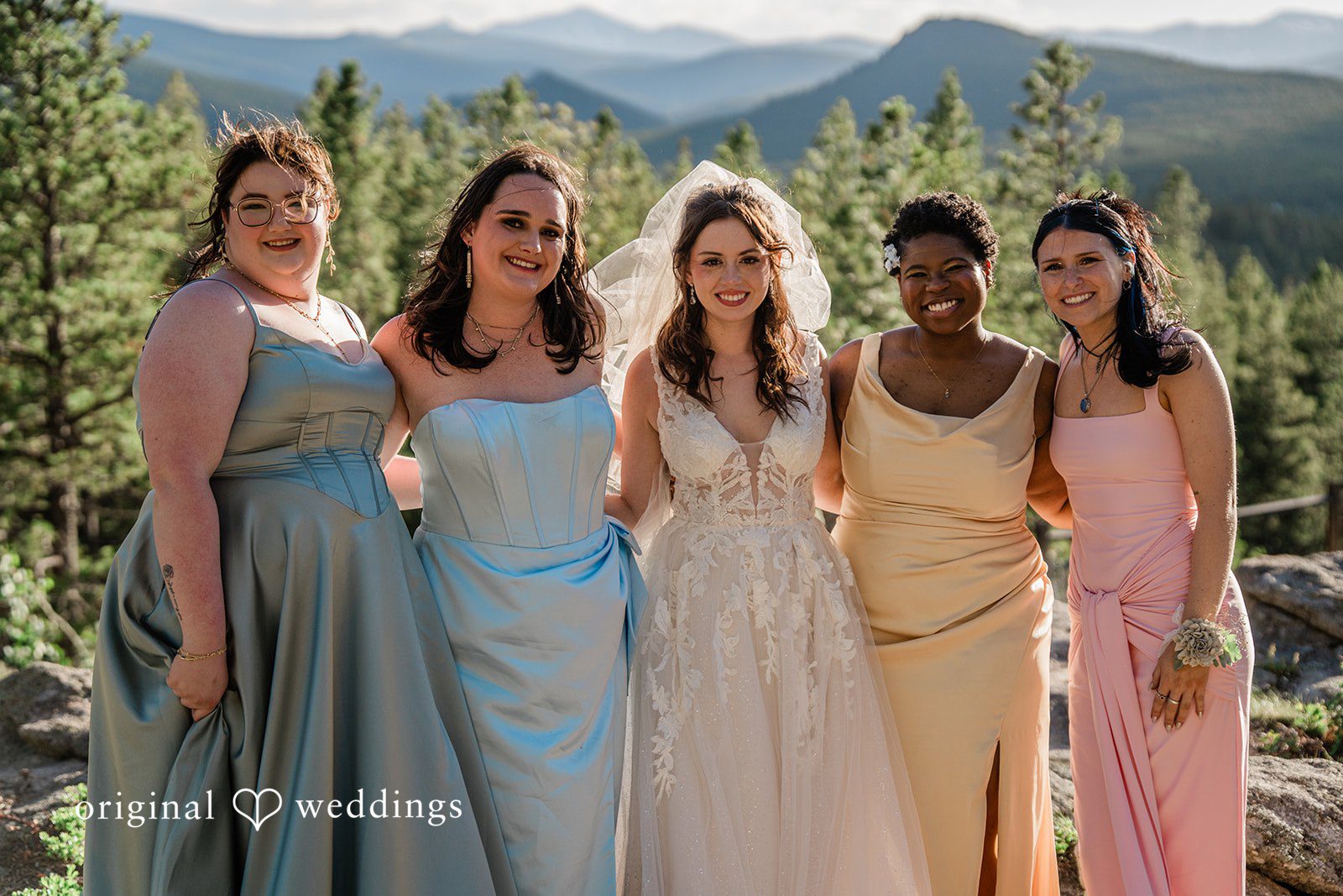 Madelyn + Erin Four bridesmaids wearing pastel dresses standing together in front of trees.
