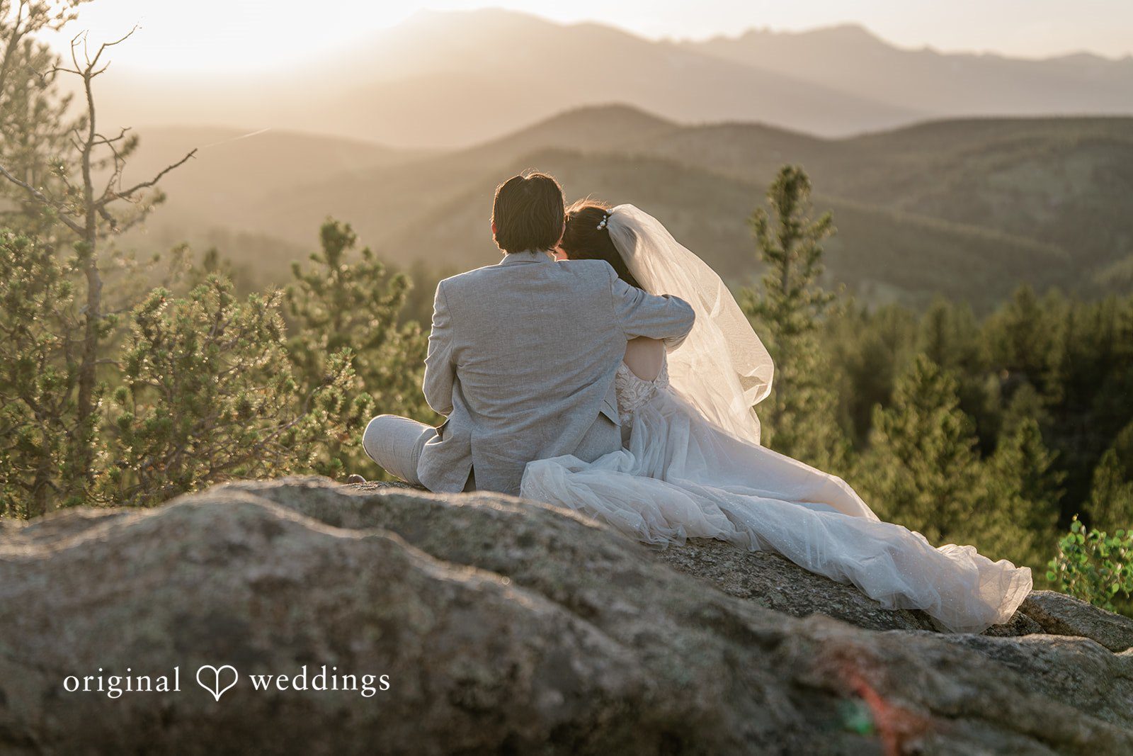 Madelyn + Erin A bride and groom sitting together on a rocky peak overlooking distant mountains.