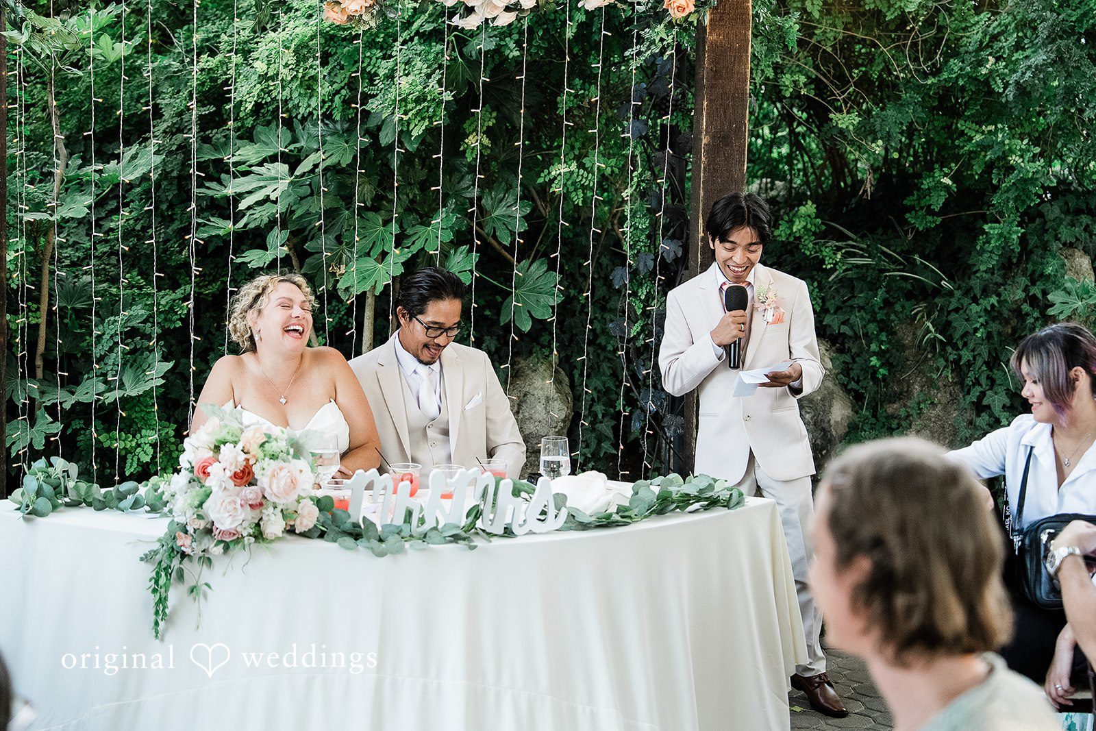Lindsay + Ryan A wedding party seated at a long reception table while someone speaks into a microphone.