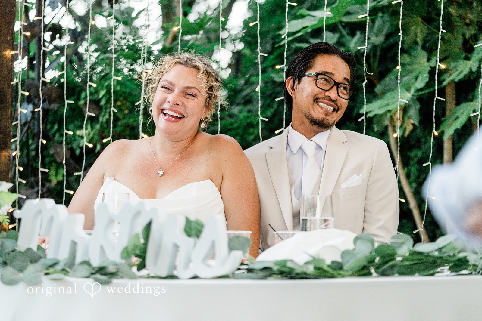 Lindsay + Ryan A bride and groom smiling happily behind a greenery-covered reception table.