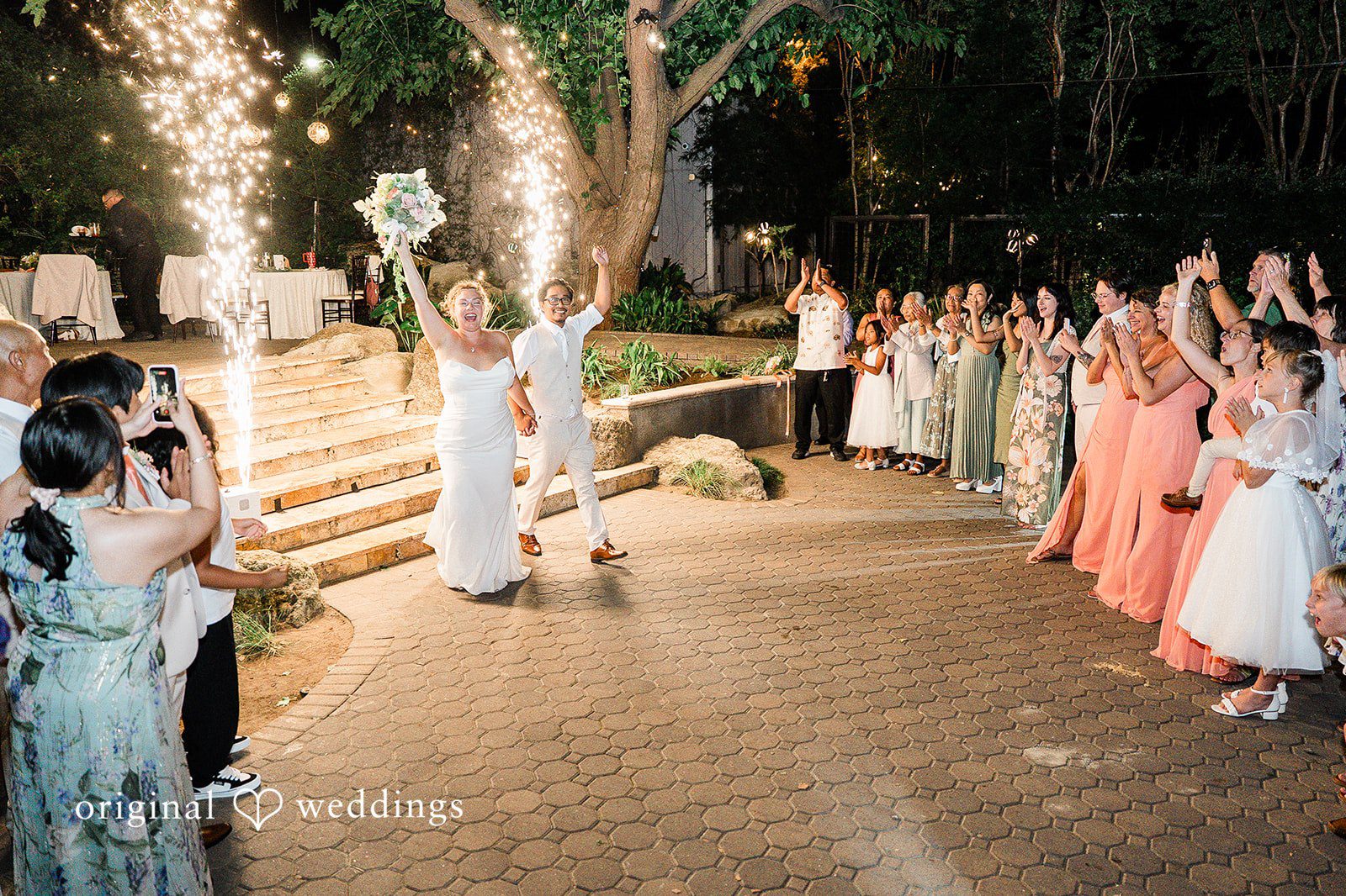 Lindsay + Ryan A bride and groom walk excitedly into their wedding reception past sparkling fountains while guests applaud.