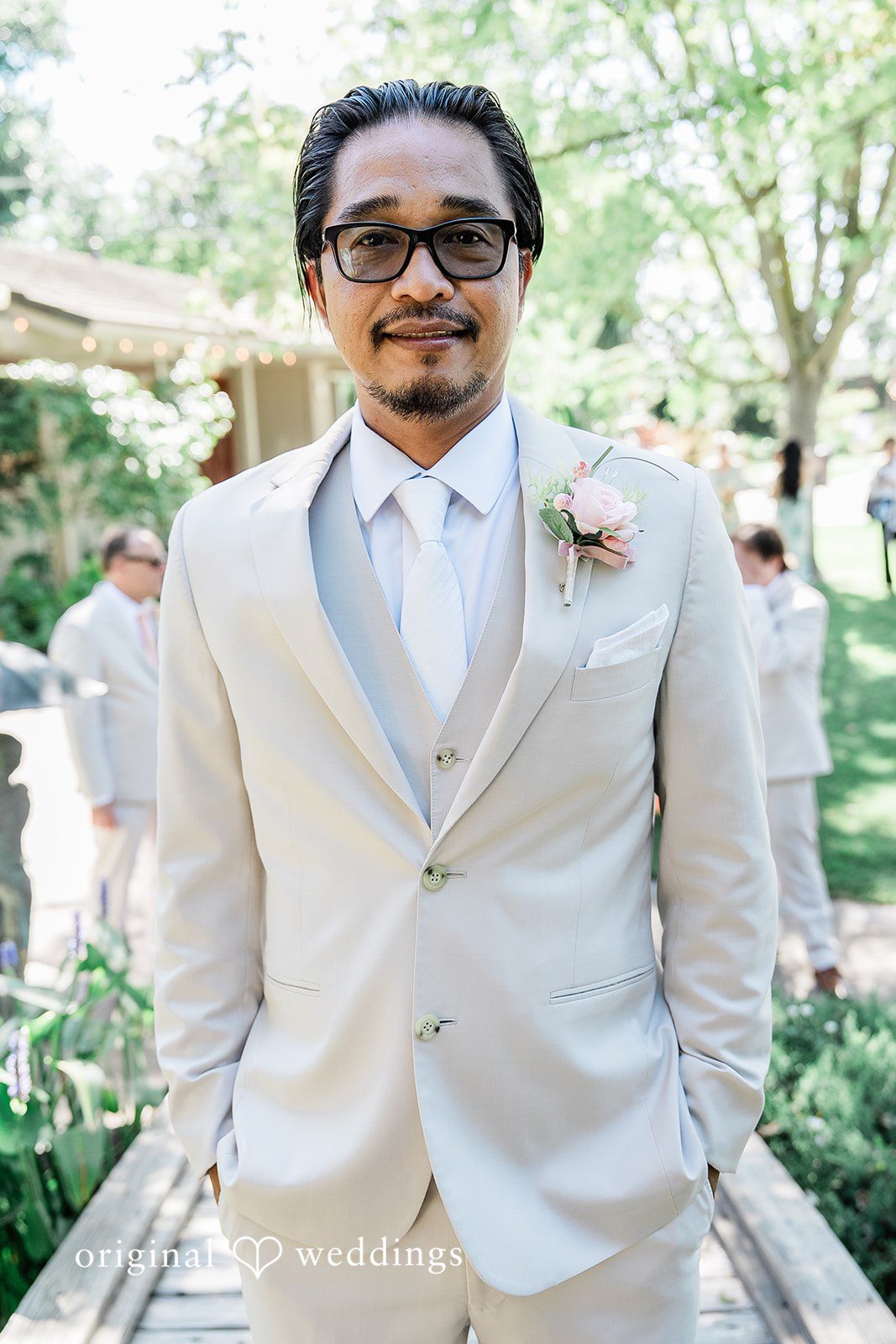 Lindsay + Ryan A groom in a light cream suit smiling warmly in a sunlit garden setting.