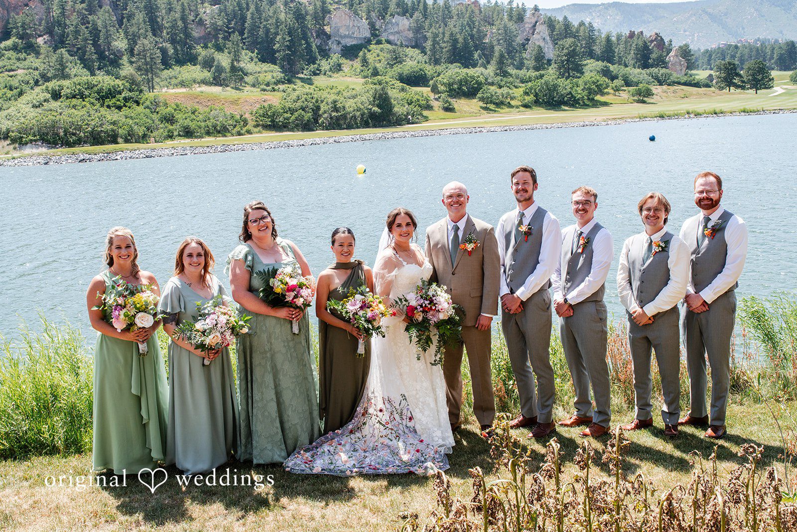 Mckenna + Ryan A large bridal party in sage green and grey attire posing together by a lake.