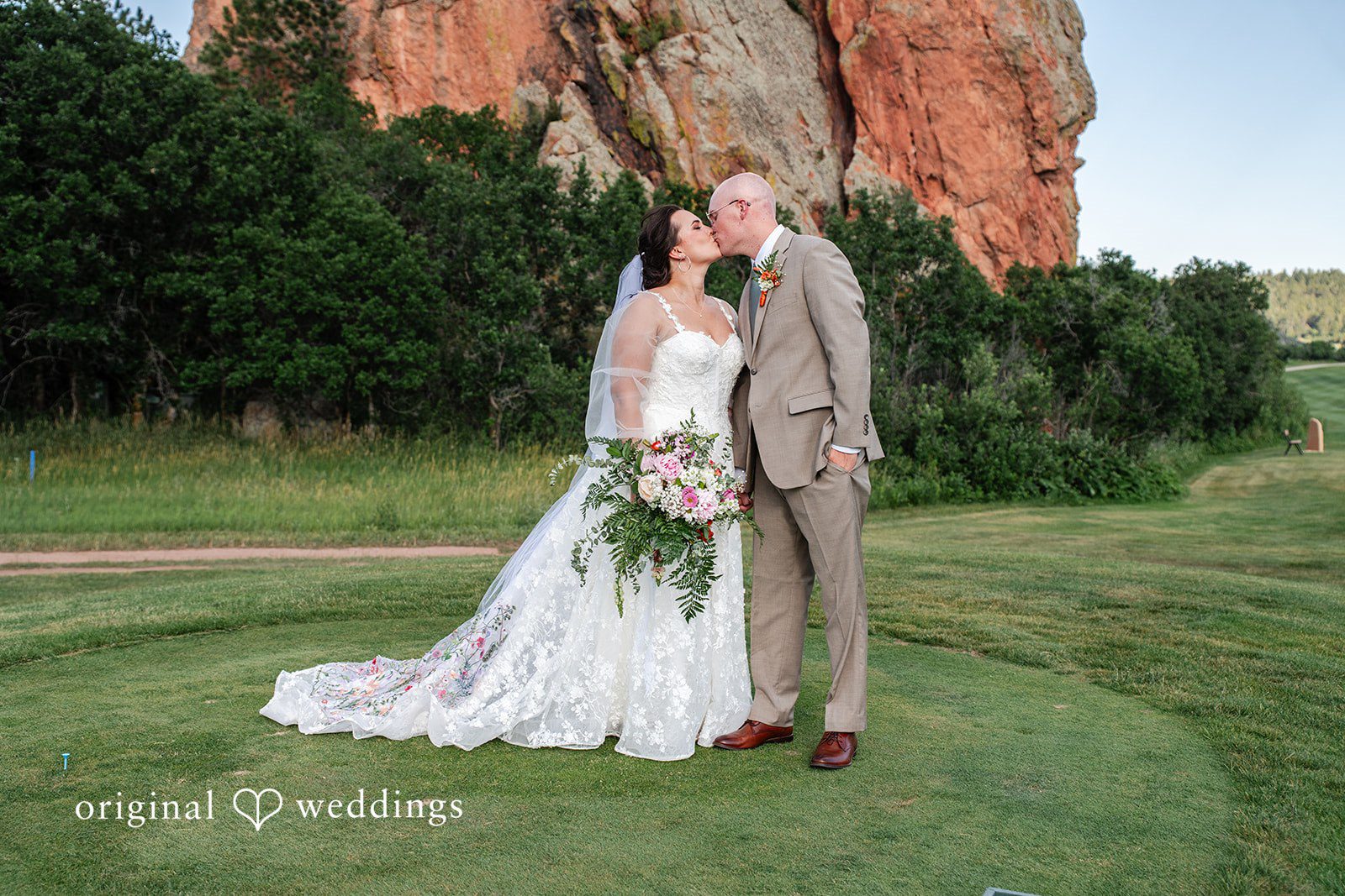 Mckenna + Ryan A bride and groom embracing on a green lawn with rocky mountains in the background.
