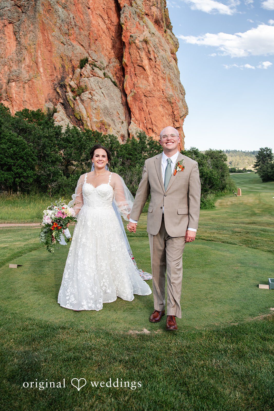 Mckenna + Ryan A bride and groom walking hand-in-hand across a grassy field near red rock formations.