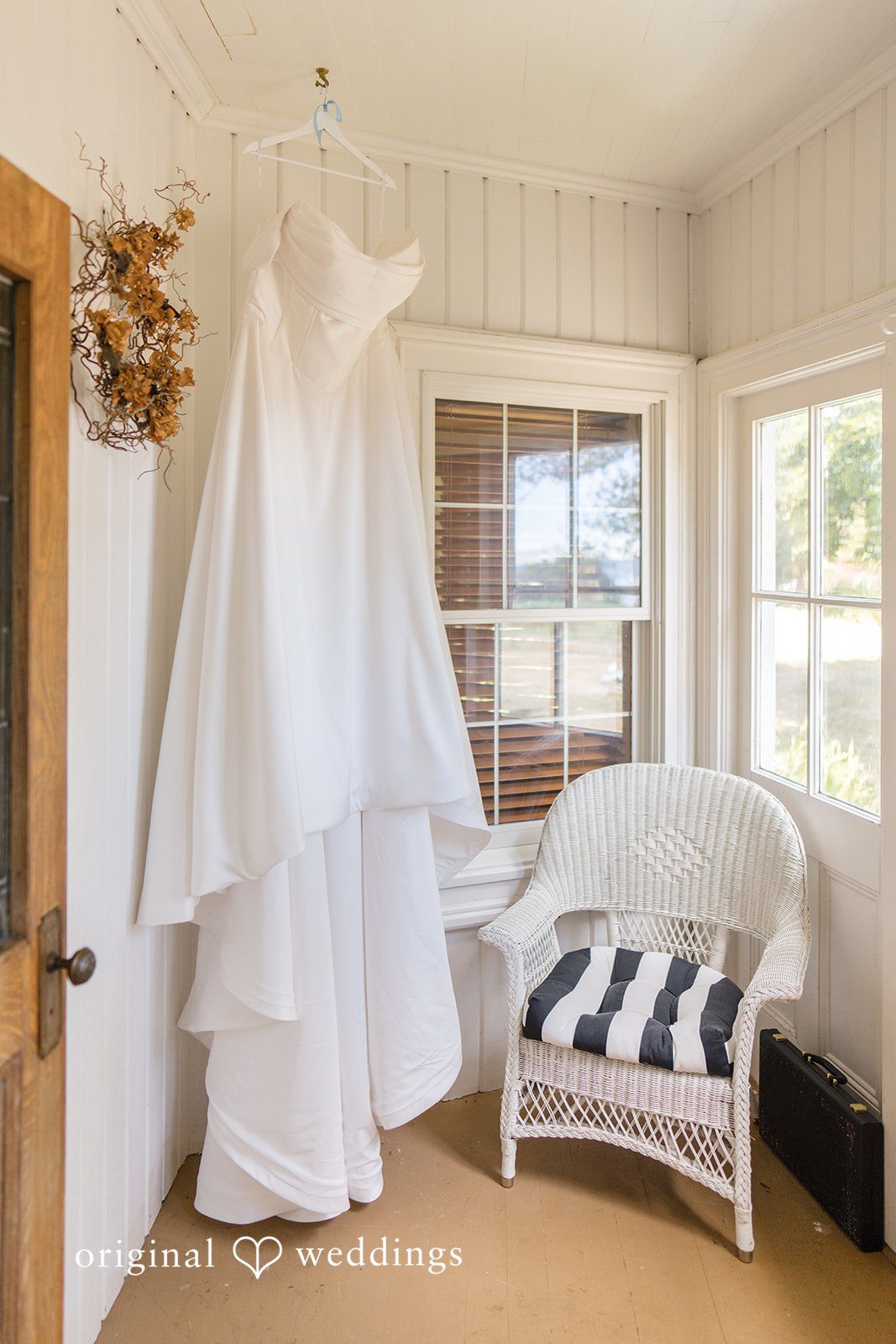 Madelyn + Connor A white wedding gown hanging by a window near a white wicker armchair.