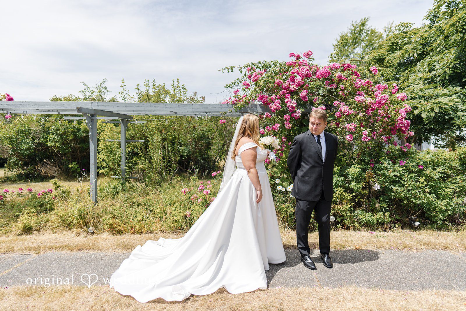 Madelyn + Connor A man in a black suit turns around to look at a bride standing before him near a pink floral bush.
