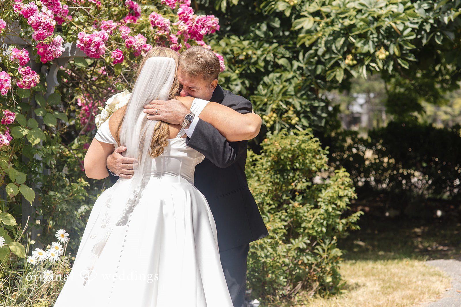 Madelyn + Connor A bride and a man in a black suit share an emotional embrace next to a blooming pink rose bush.