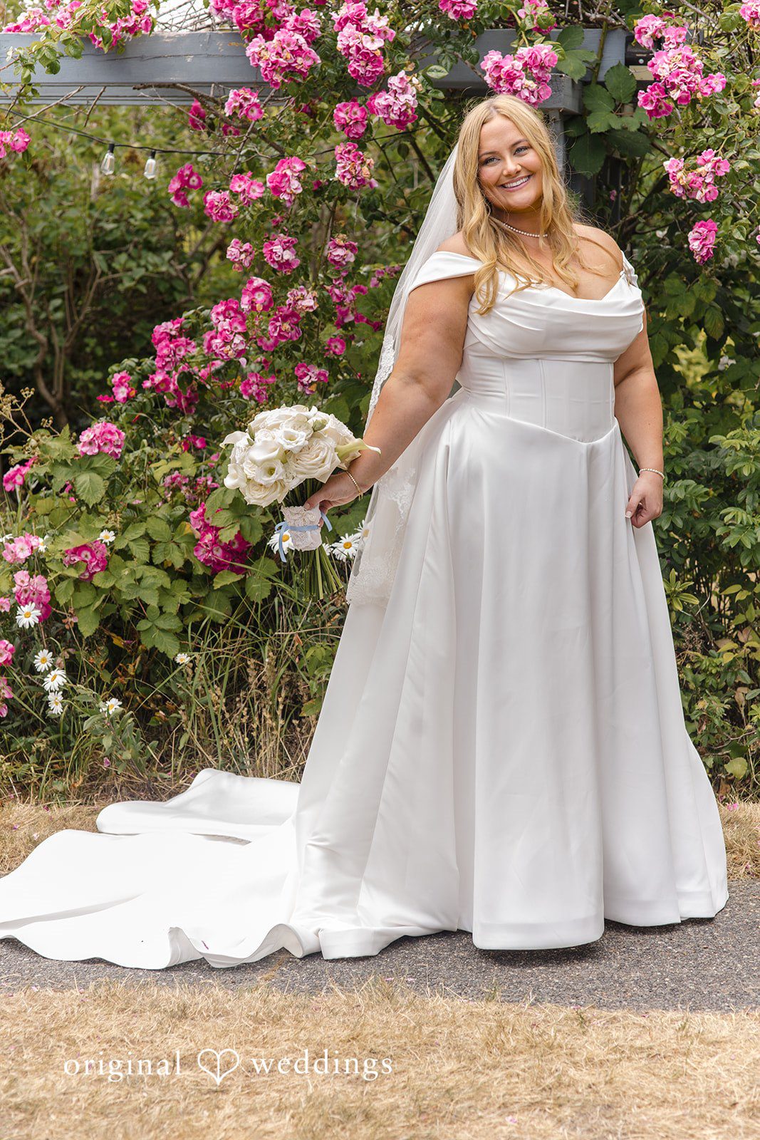 Madelyn + Connor A bride posing in front of a green bush filled with pink flowers.