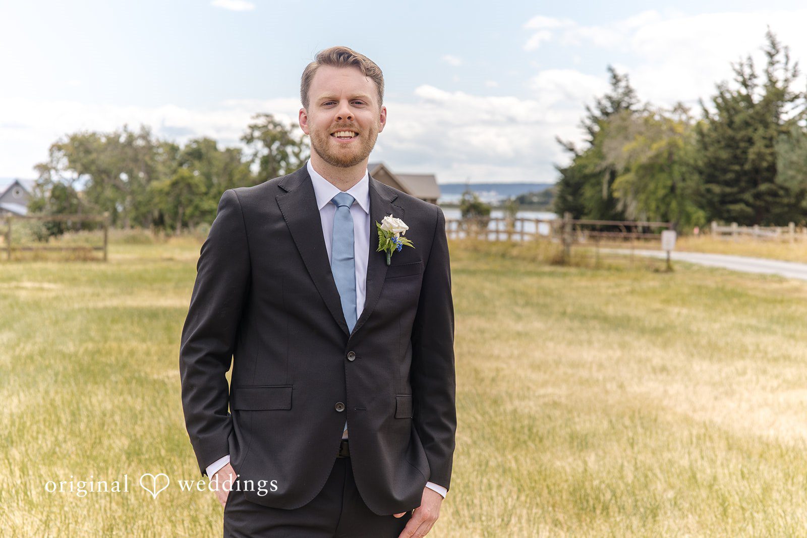 Madelyn + Connor A groom in a dark suit smiling while standing on a path in a field.