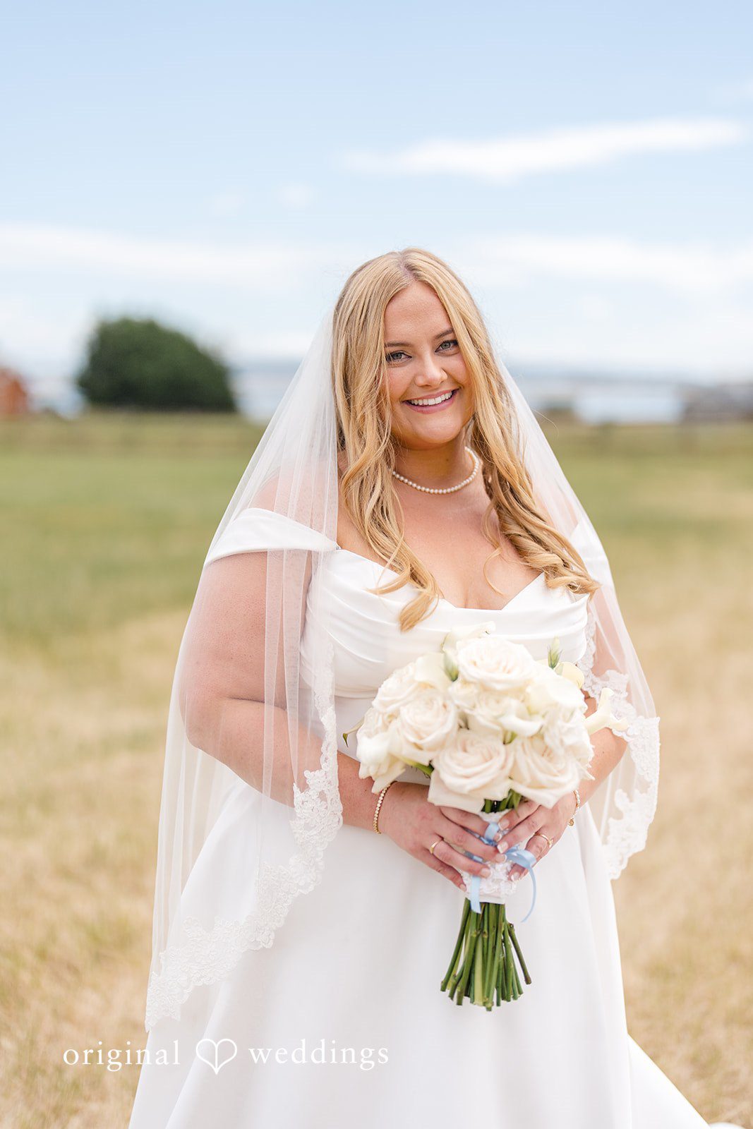 Madelyn + Connor A smiling bride holding a white bouquet while posing in an open grassy area.