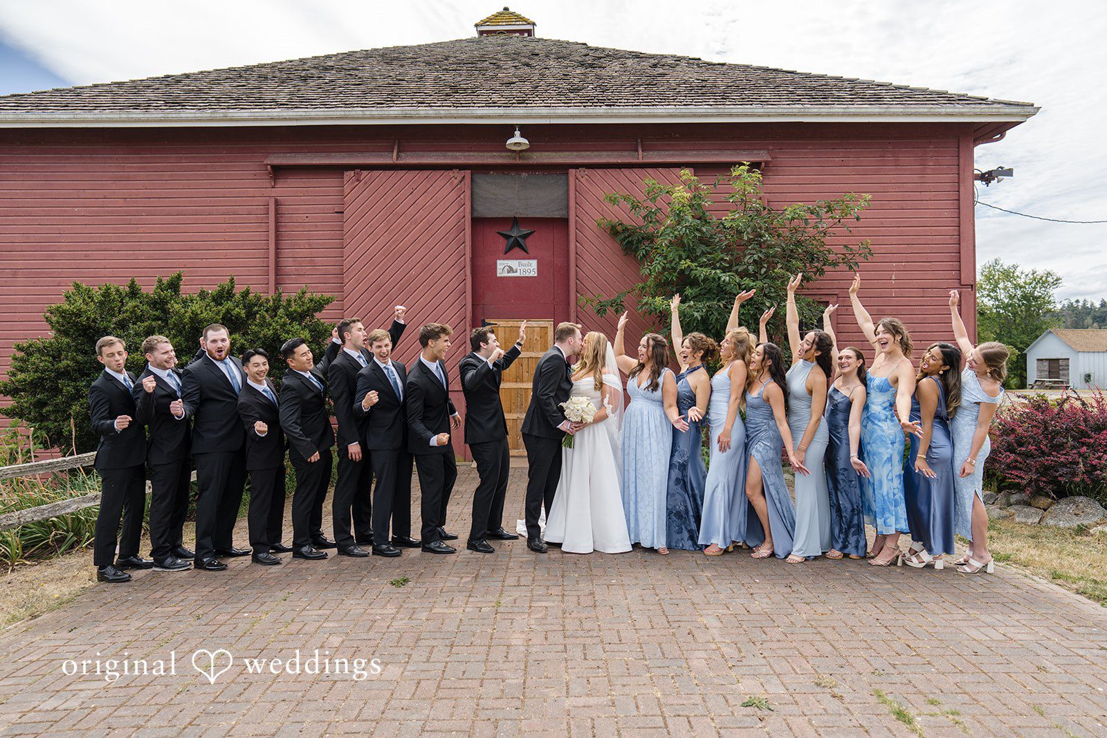 Madelyn + Connor A full wedding party with bridesmaids and groomsmen posing in front of a red building.