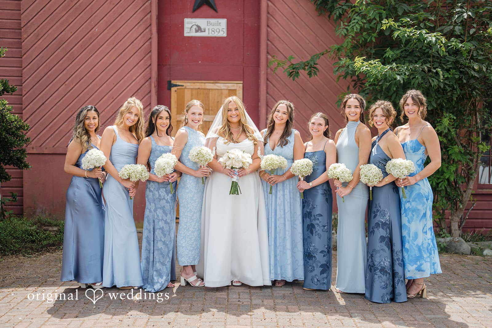 Madelyn + Connor Bride and bridesmaids dressed in different shades of blue dresses posing together by a red building.