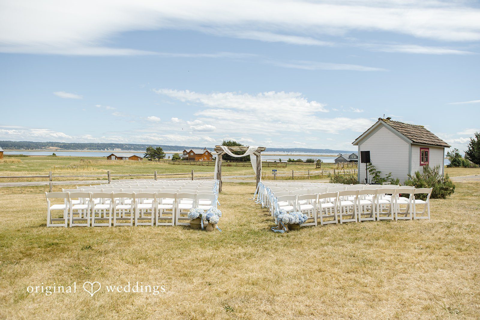 Madelyn + Connor An empty ceremony setup with rows of white chairs facing a wooden arch in an open field.