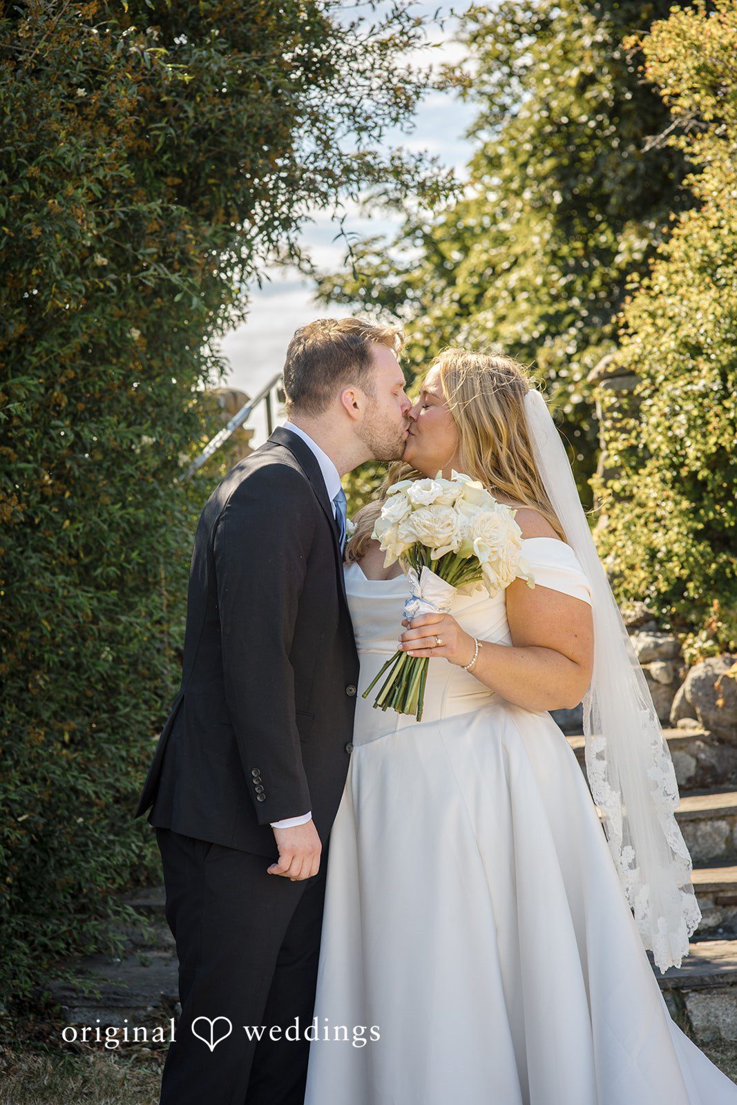 Madelyn + Connor A bride and groom sharing a kiss against a background of lush green trees.