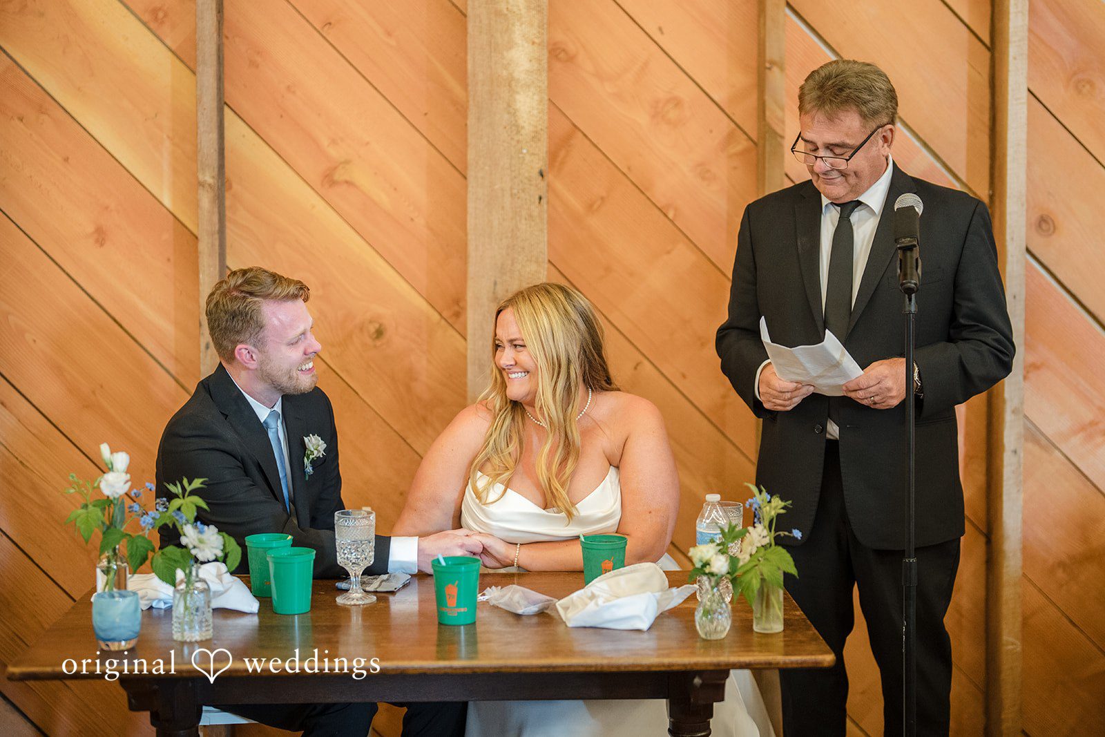 Madelyn + Connor A man in a suit speaking into a microphone next to a seated bride and groom at a reception table.